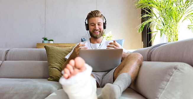 Young man with a broken leg in a cast working on a laptop on the sofa