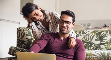 Woman sitting on sofa with one arm on man sat on floor looking at a laptop